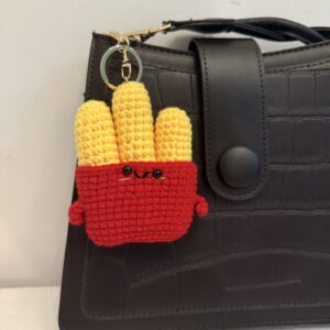 Rot-gelber gehäkelter Pommes-Schlüsselanhänger mit Gesicht und goldfarbenem Ring, an einer dunklen Tasche befestigt.
