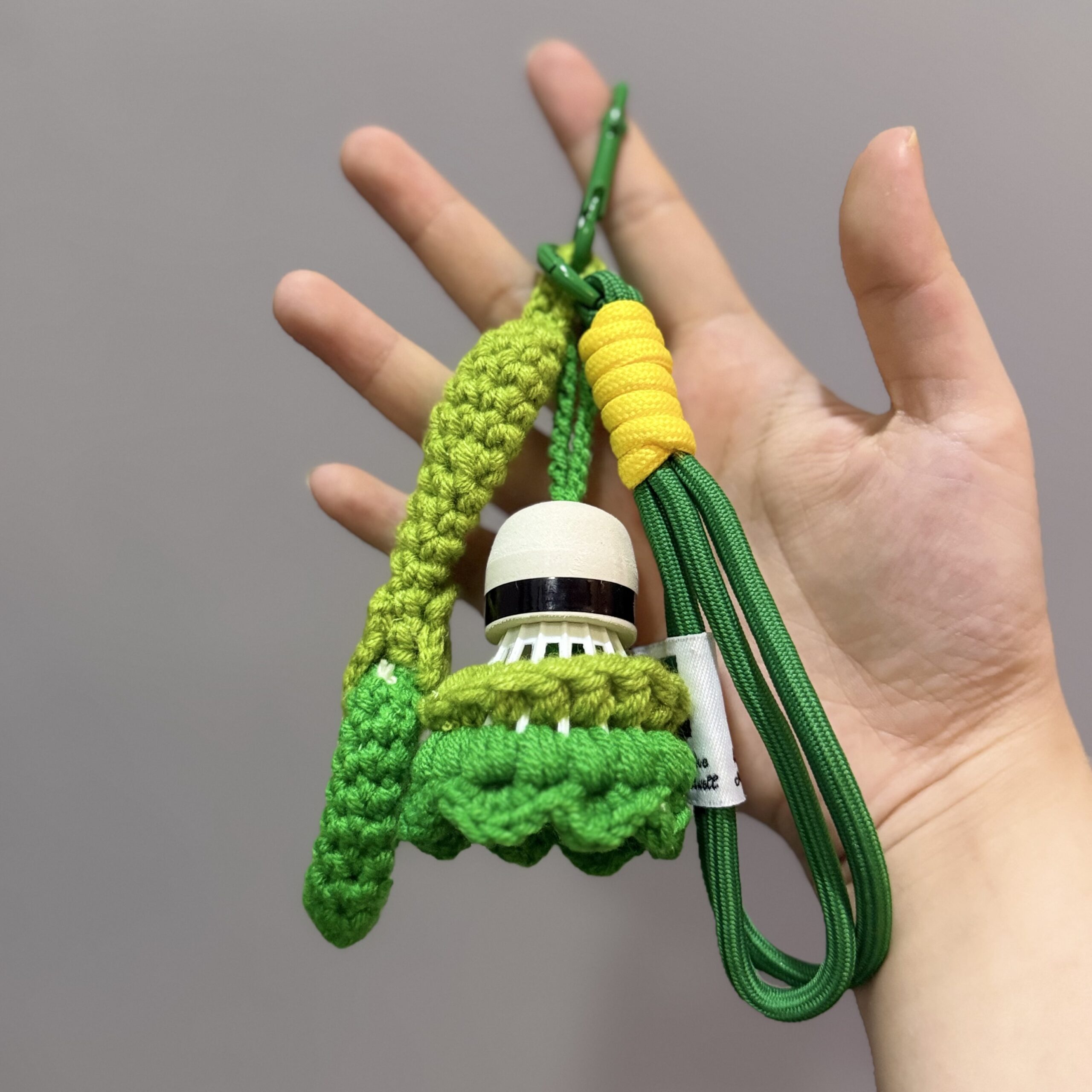 Grüner handgemachter Federball-Schlüsselanhänger mit Schläger-Motiv, gelbem Kordeldetail und grünem Karabiner in der Hand.