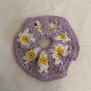 Lila handgemachter Scrunchie mit weißen Gänseblümchen, gelben Blütenmitten und weicher Maschenstruktur auf hellem Stoff.