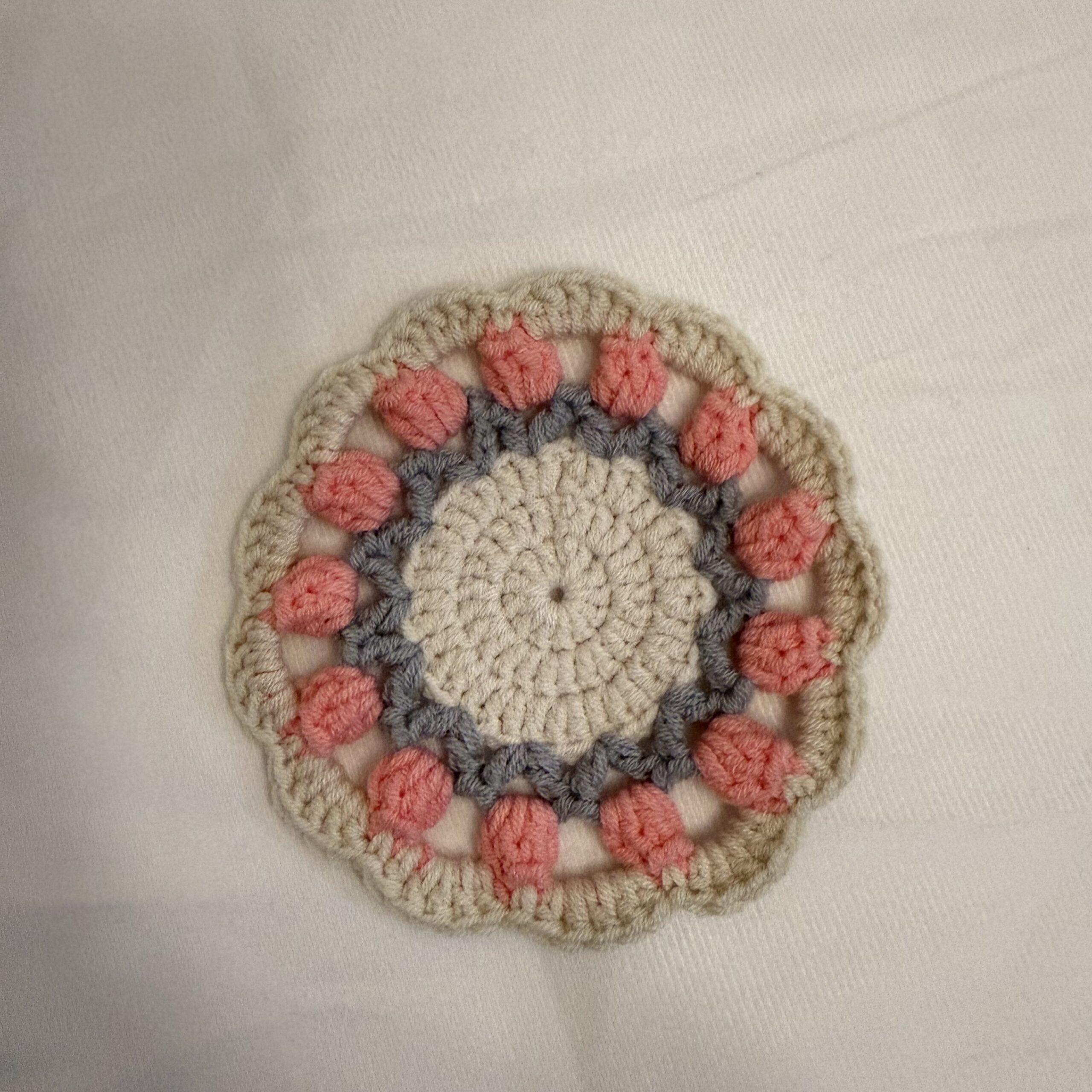 Runder handgemachter Tassenuntersetzer mit rosa Tulpenmuster, grauem Ring, cremefarbener Mitte und gewelltem Rand auf hellem Stoff.