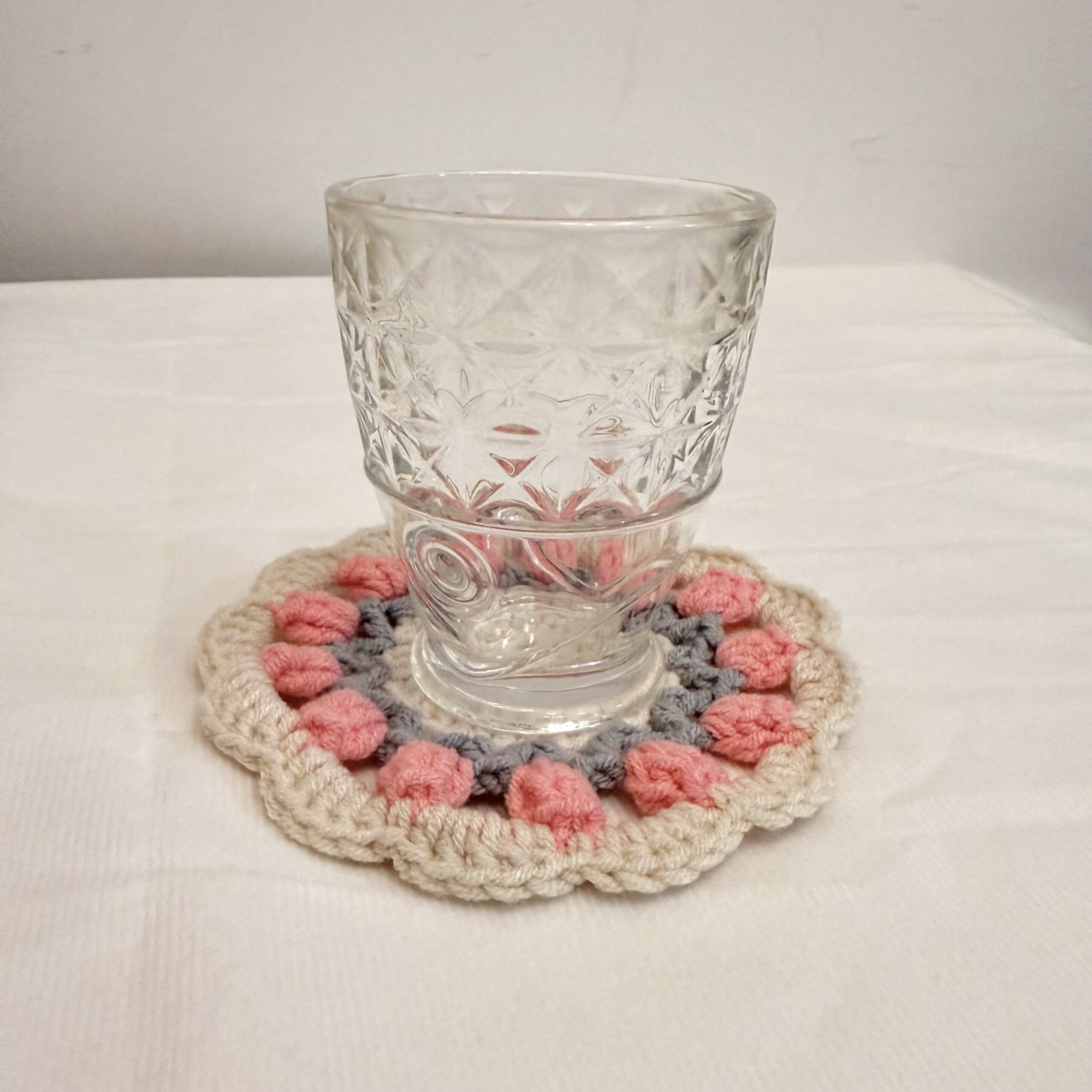 Glas auf rosa Tulpen-Tassenuntersetzer, handgemachte Maschenstruktur und cremefarbener Rand seitlich sichtbar.