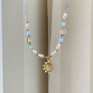Hellblaue Perlenkette mit weißen Kunstperlen, goldfarbenen Zwischenperlen und Sonnenanhänger vor hellem Vorhang.