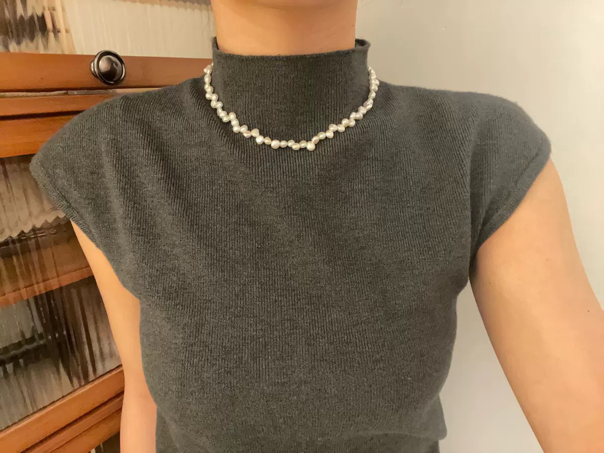 Weiße Perlenkette mit unregelmäßigen Perlen als kurze Schlüsselbein-Kette getragen zu einem grauen Oberteil.