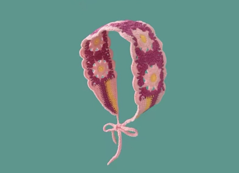 Rosa Häkel-Haarband mit Gänseblümchen-Muster und Bindebändern vor türkisfarbenem Hintergrund.