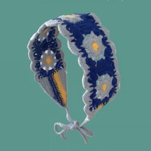 Gebogenes blaues Strick-Haarband mit hellblauen Gänseblümchen und gelben Blütenmitten vor türkisfarbenem Hintergrund.