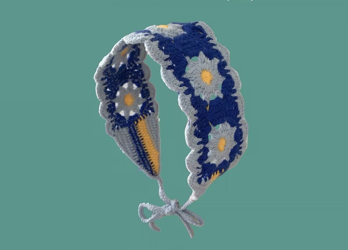 Gebogenes blaues Strick-Haarband mit hellblauen Gänseblümchen und gelben Blütenmitten vor türkisfarbenem Hintergrund.