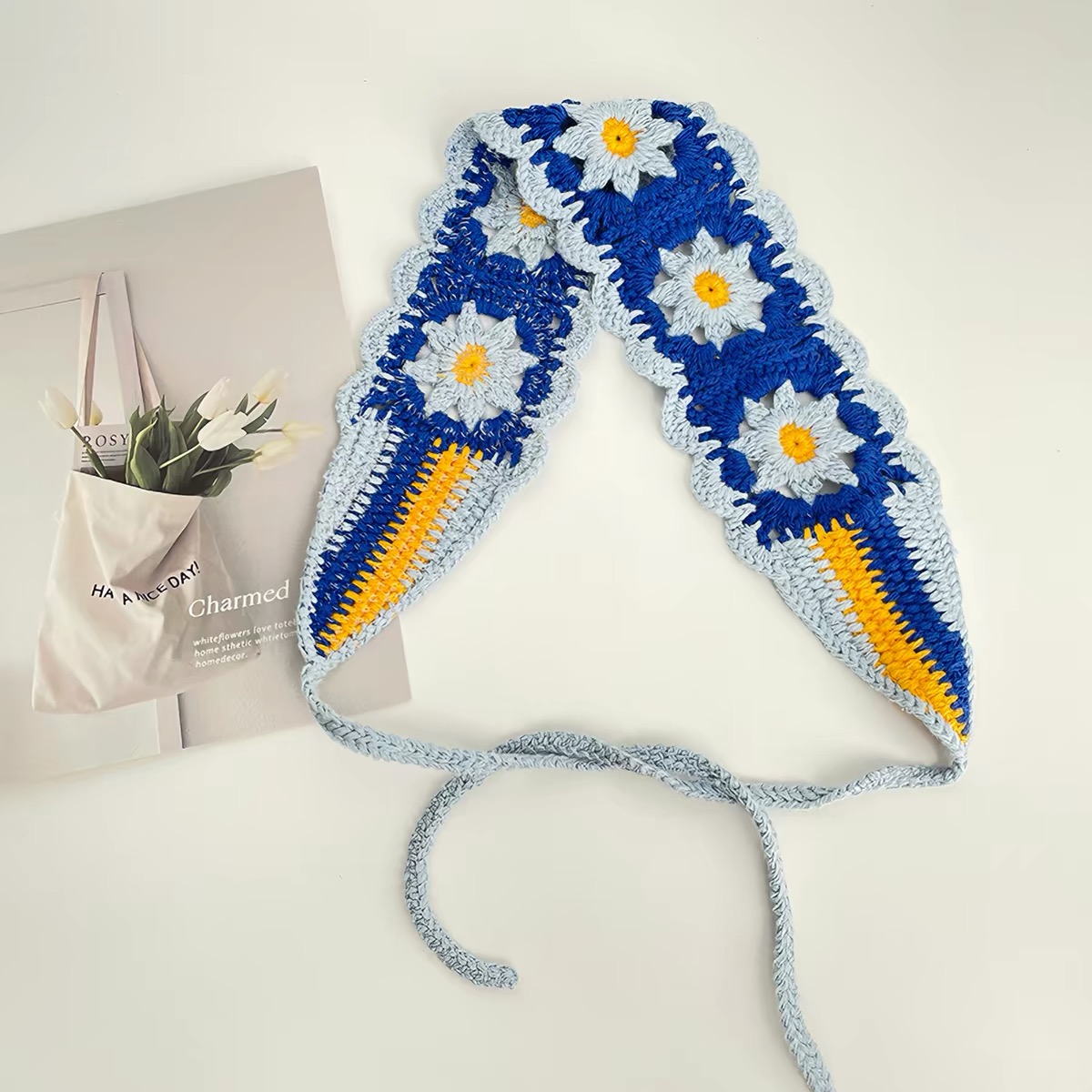Blaues Strick-Haarband mit floralen Motiven, gelb-blauen Endstreifen und Bindebändern auf hellem Hintergrund mit dekorativer Karte.