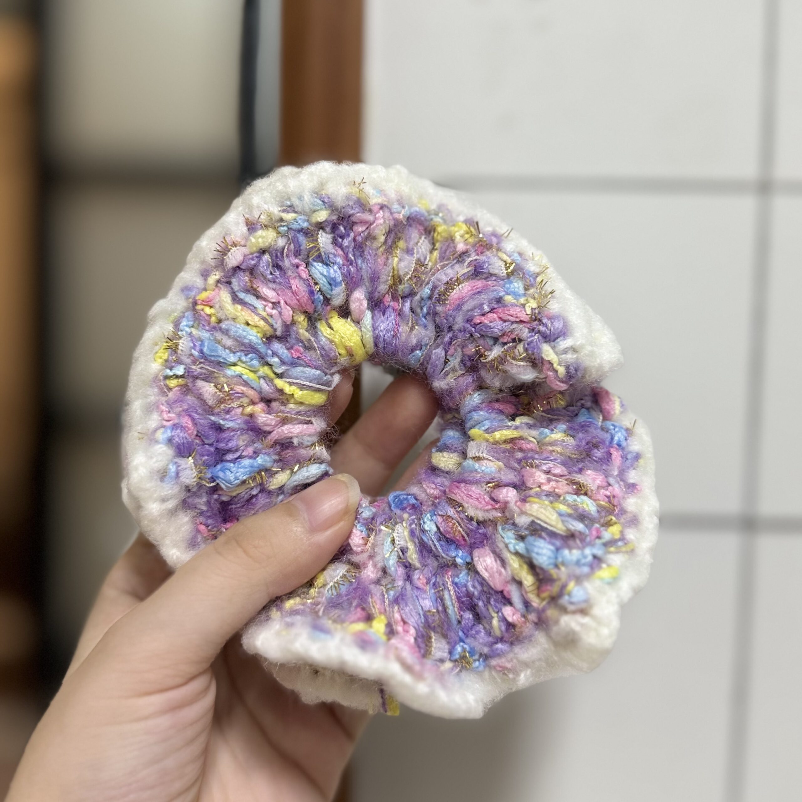 Nahaufnahme eines lila bunten Scrunchies mit weißem welligem Rand, rosa, blauen und gelben Garnakzenten
