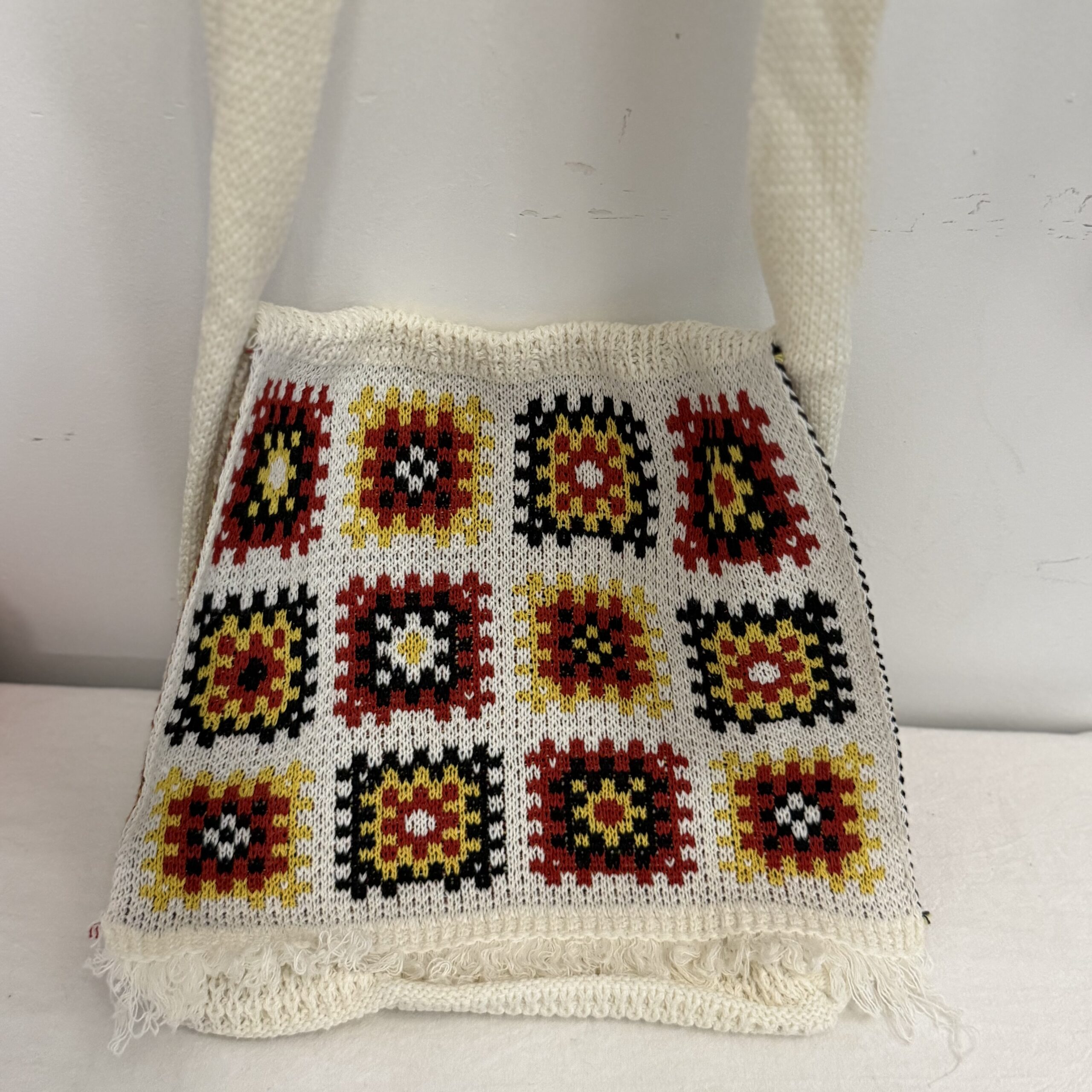 Nahaufnahme einer weißen Granny-Square-Stricktasche mit geometrischem Muster, breiten Trägern und fransiger Unterkante.