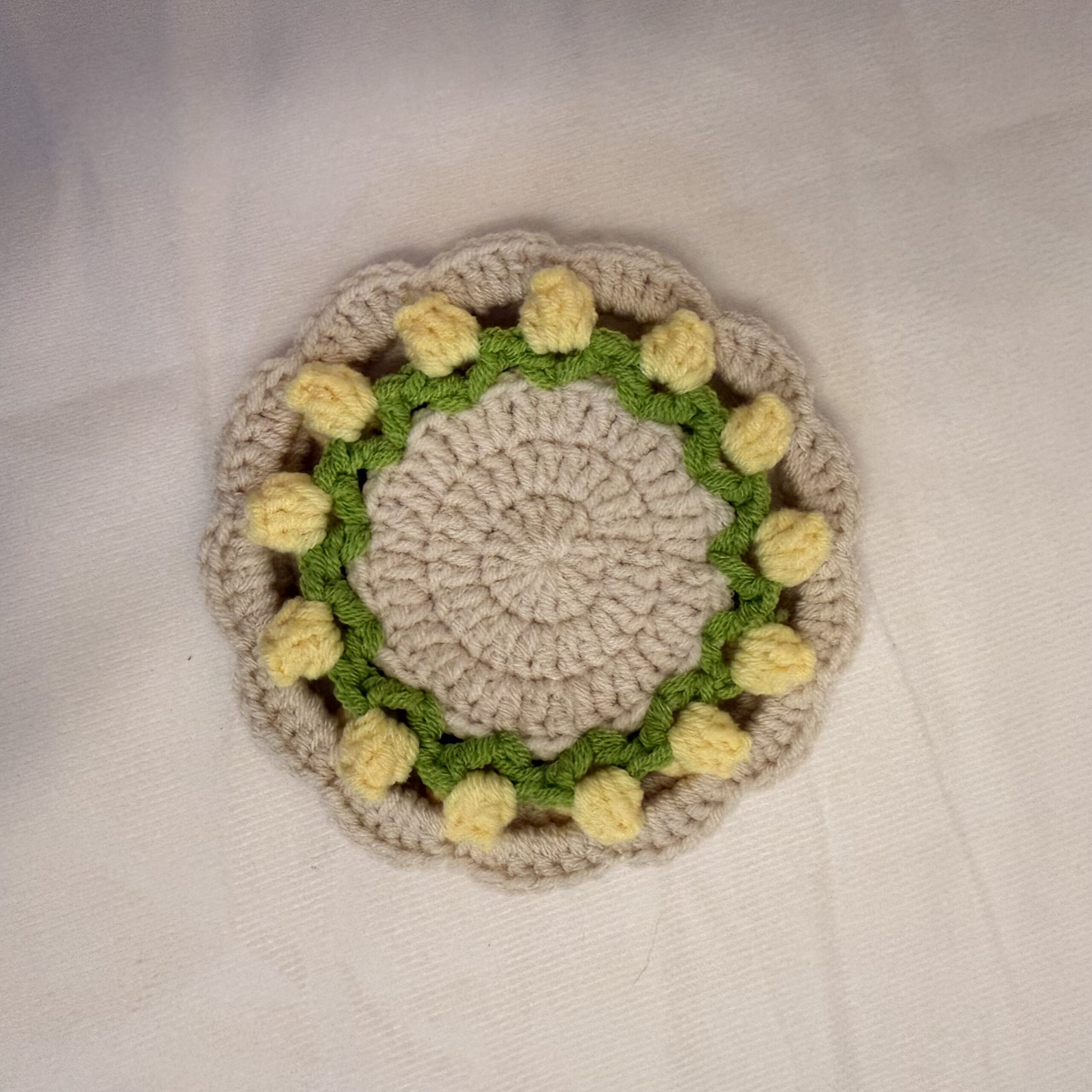 Runder handgemachter Tassenuntersetzer mit gelben Tulpen, grünem Ring, cremefarbener Mitte und gewelltem Rand auf hellem Stoff.