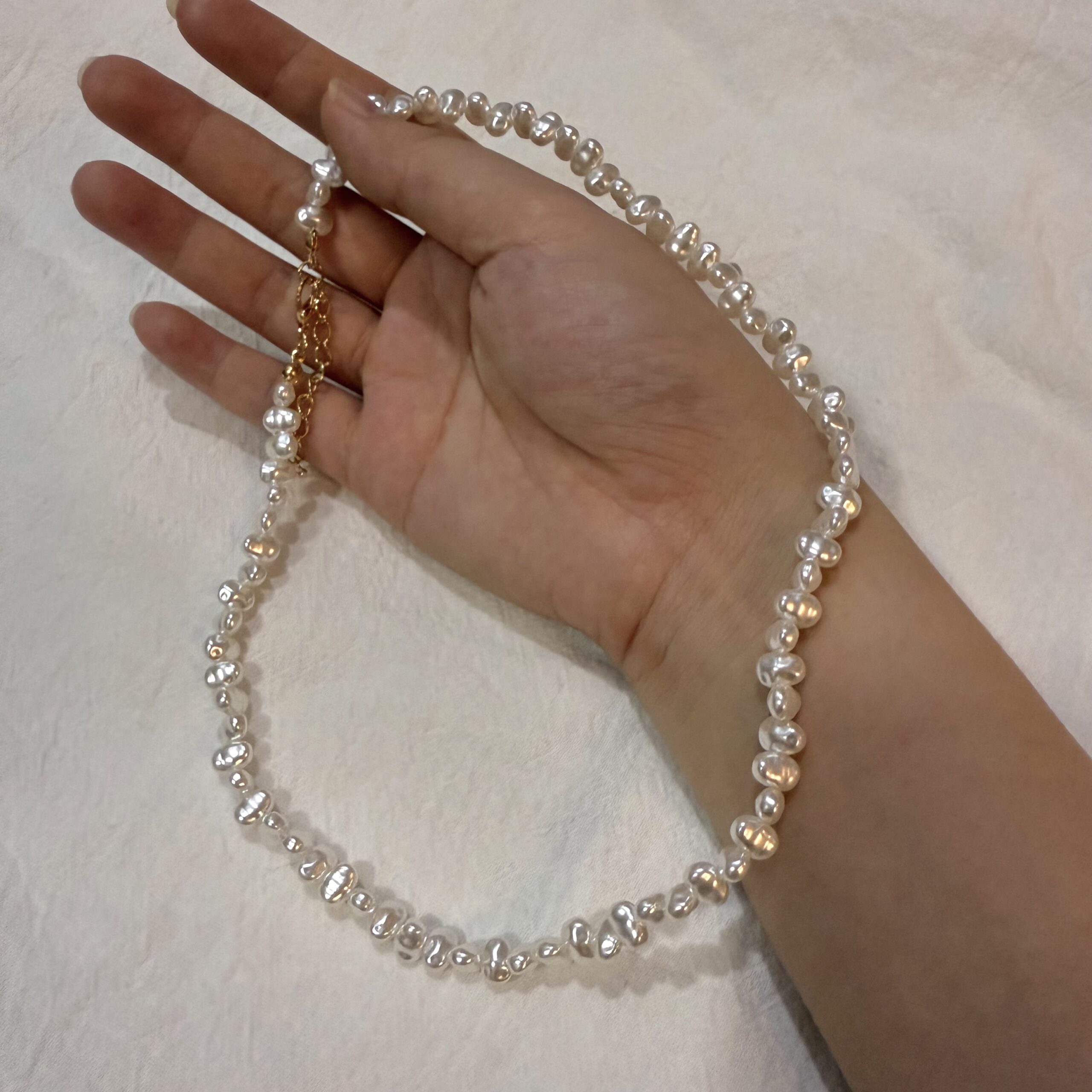Nahaufnahme einer weißen Perlenkette in der Hand mit schimmernden unregelmäßigen Perlen und goldfarbenem Verschluss.